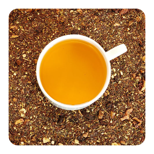 A cup of tea on a bed of dried honeybush and rooibos tea leaves with hazelnut, apple, and raisin pieces. 