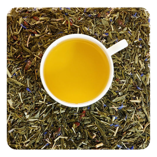 A cup of pomegranate green tea served on a bed of dried green tea leaves with visible pieces of fruit and flower petals, indicating the ingredients of the tea blend.